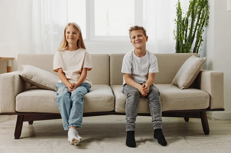 Smiling boy and girl sitting on beige couch in modern bright living room, casual clothes, natural light, relaxed atmosphere, home interior, children portrait concept.