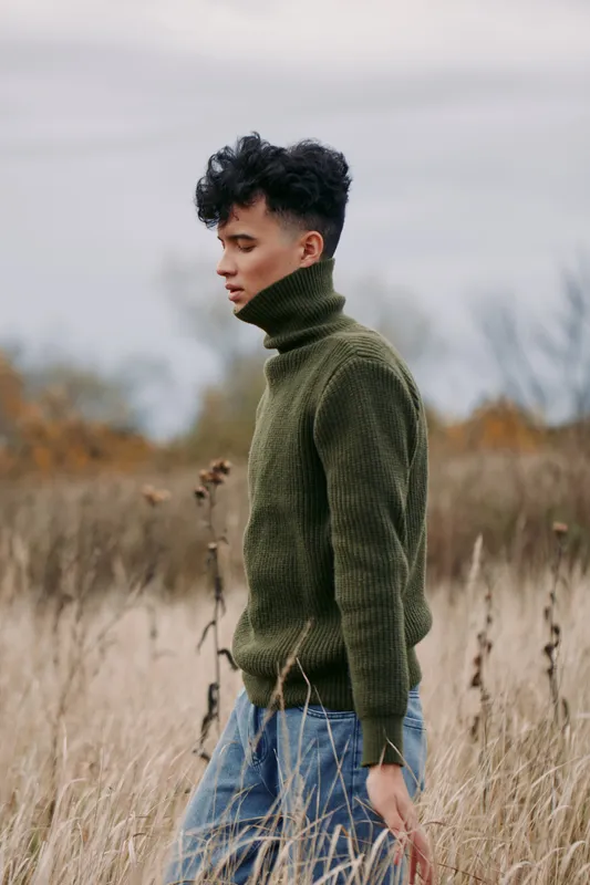 A contemplative young man in a green turtleneck walks through tall dry grass, natural light and muted palette conveying quiet reflection and a sense of authenticity and credibility in the scene.