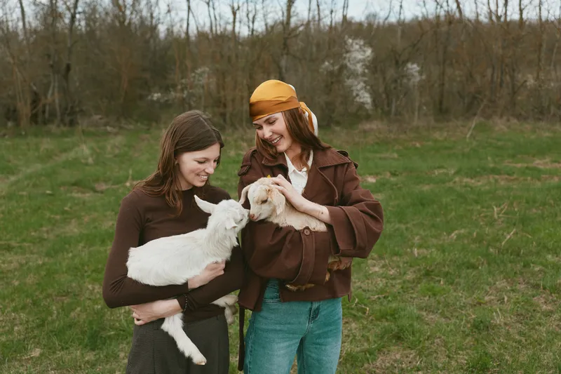 Two women in casual outdoor clothing smile while holding a goat kid in a sunlit field, capturing everyday bond, compassion, and gentle animal care in a warm countryside moment.