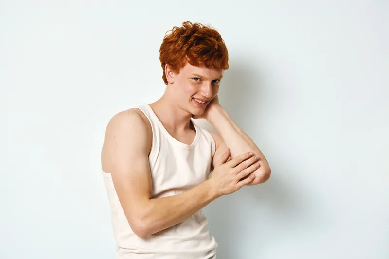 Young man with red hair wearing white tank top posing and smiling against white background indoor, casual style and confident expression for portrait photo.