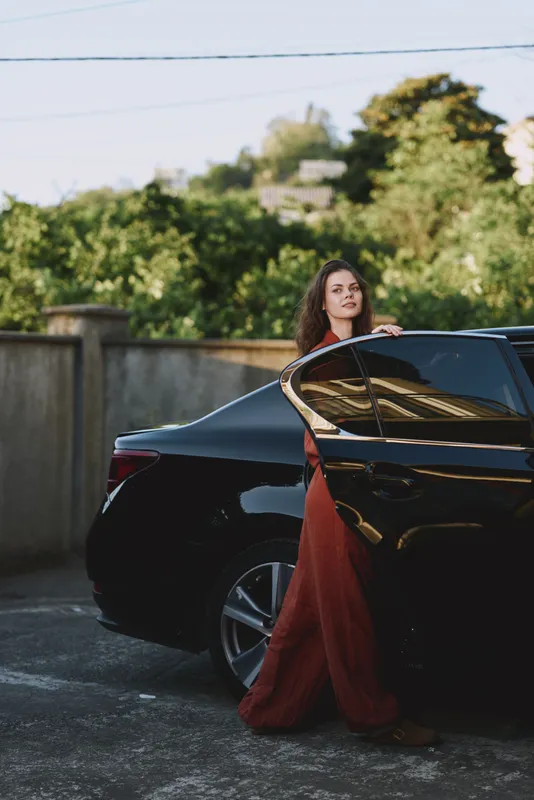 A stylish woman in a flowing red gown leans against a sleek black car in a sunny driveway, capturing a bold lifestyle moment with confident poise and cinematic lighting