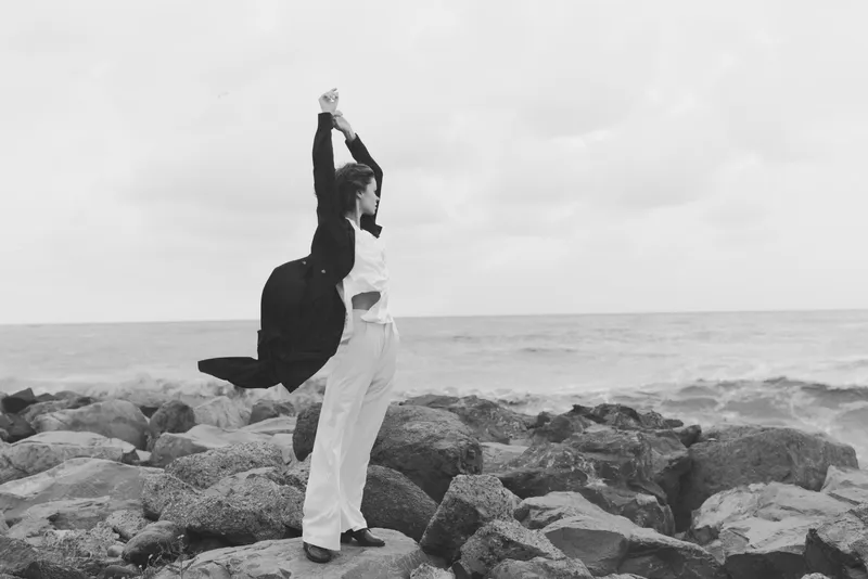 Motion of a figure leaping on rocky shore beside the sea, wind in clothing, black and white capture of outdoor freedom and balance between water and stone in a dramatic, graceful frame that