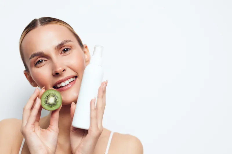 Natural glow and relaxed smile as a young woman with blonde hair holds a kiwi near her face and a white cosmetic bottle, presenting a fresh skincare moment in a bright minimal setting.