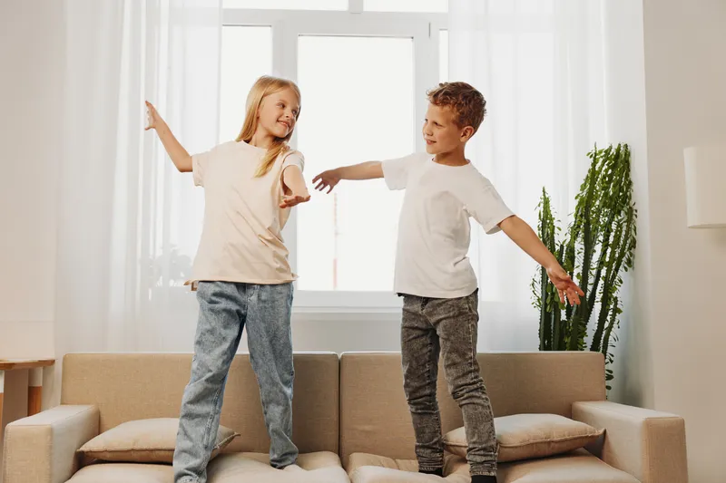 Two happy children standing on a sofa with arms outstretched in a bright living room. Casual clothes and natural light create a playful and joyful family moment indoors.