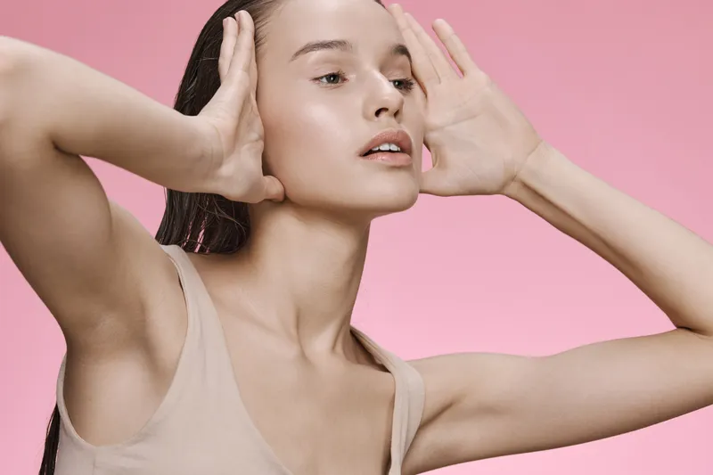 A young female model poses gracefully against a soft pink background, showcasing natural beauty with a serene expression and minimal makeup, emphasizing healthy skin