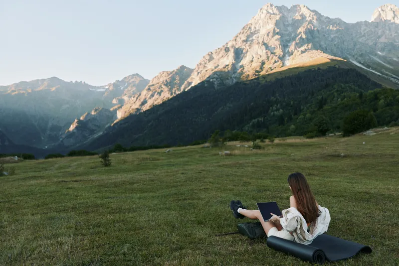 A solitary worker sits on a mat in a wide meadow, laptop in lap, while towering mountains rise nearby, creating a calm, productive outdoor work scene.