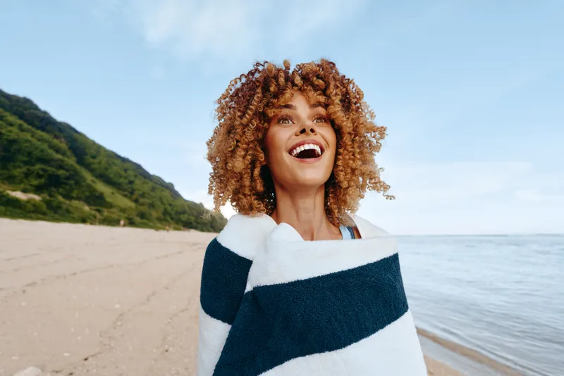 Young woman with curly hair laughs on a sunny beach wrapped in a navy and white striped towel, with blue sky and calm sea behind, creating a carefree bright moment