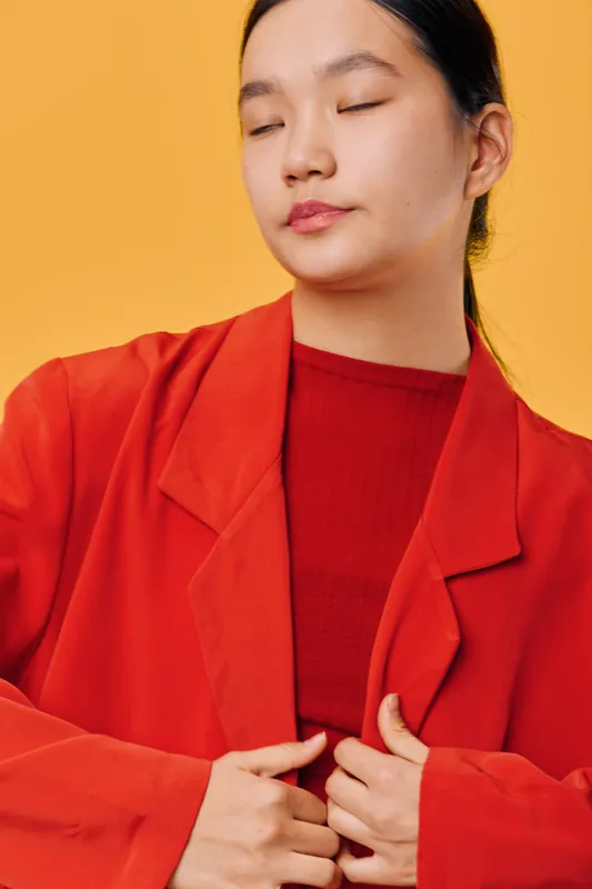Young woman with closed eyes wearing red blazer and top standing against yellow background. Portrait of calm female with dark hair styled back in minimal natural makeup.