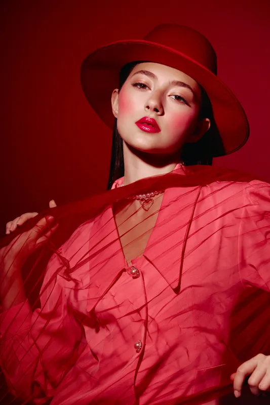Young woman dressed in a red shirt and hat strikes a confident pose with dramatic angles of sheer fabric in a creative fashion portrait against a bold red background.