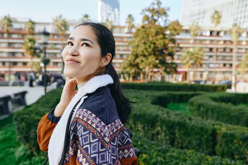 Woman in a patterned coat outdoors in a park, looking up with a gentle smile, surrounded by greenery and urban backdrop, showcasing style, autumn colors, and carefree mood.
