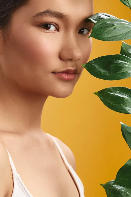 Asian woman gazes softly at the camera with a calm, confident expression. A yellow backdrop and green leaves frame her beauty, creating a fresh, natural portrait for branding.