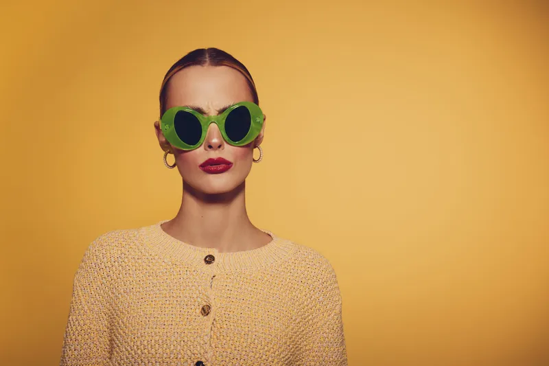 Stylish woman with slicked back hair wears large green sunglasses and yellow knitted sweater against yellow background. Fashion portrait with bold makeup and confident expression in studio lighting.