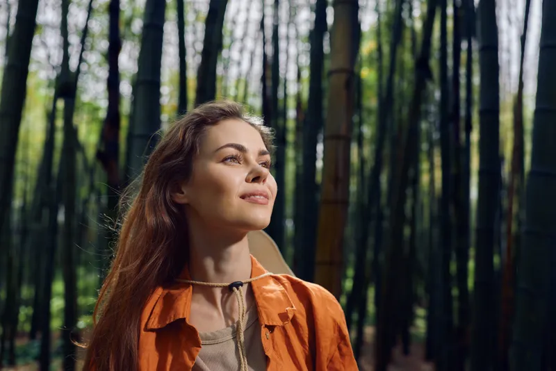 Woman portrait in bamboo forest nature outdoors with sunlight on her face, smiling young female wearing orange jacket, serene expression and peaceful gaze among tall green trees.