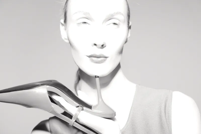 Caucasian woman with pale skin holds high heel shoe near face, eyes closed, wearing sleeveless top. Concept of fashion, elegance, and beauty with minimalistic style.