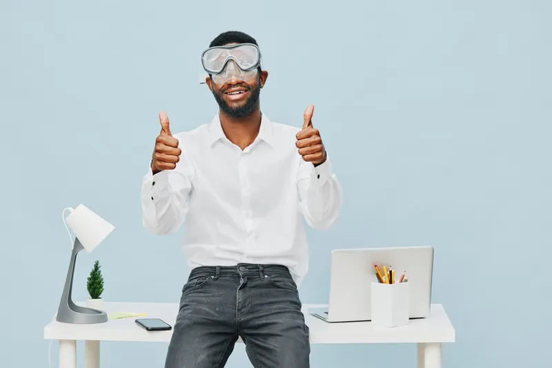 Smiling man with glasses giving thumbs up while standing at office desk, wearing white shirt, dark jeans, and safety goggles, isolated on light blue background. Happy work environment concept, positiv