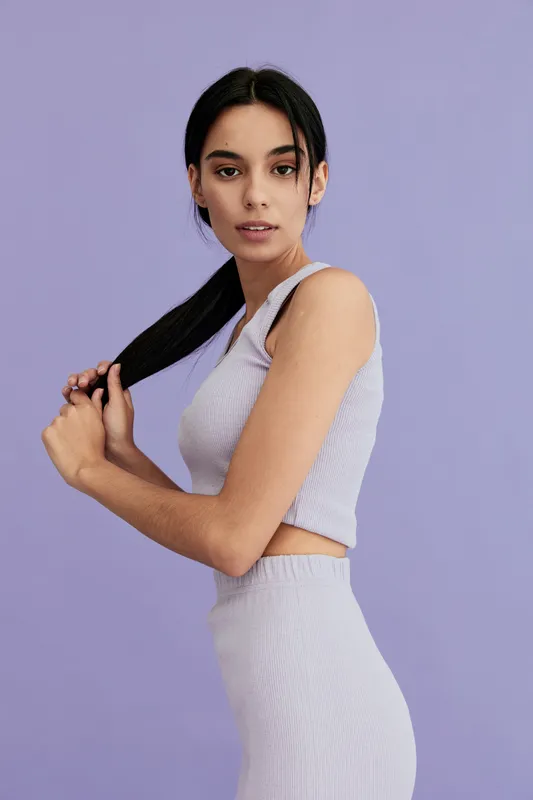 Young woman with dark hair wearing a light purple sleeveless outfit stands against a violet background, holding her ponytail confidently and looking into the camera.