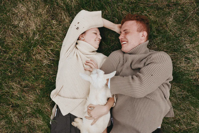 Cheerful couple outdoors, lying on grass with a playful goat kid between them, cozy sweaters, warm sunlight, candid moment, relaxed embrace, natural tones, countryside vibe