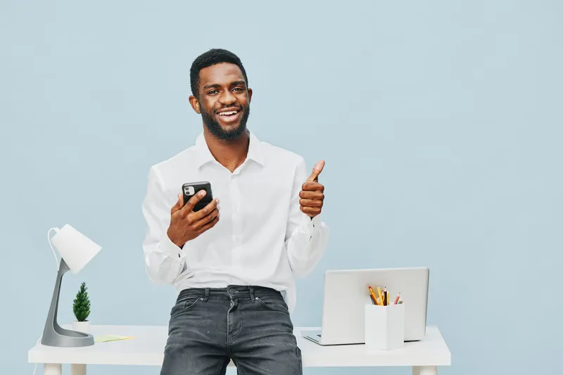 Confident smiling man wearing white shirt holding smartphone, gesturing thumbs up, standing at modern workspace with desk lamp and laptop, light blue background, business and technology concept.