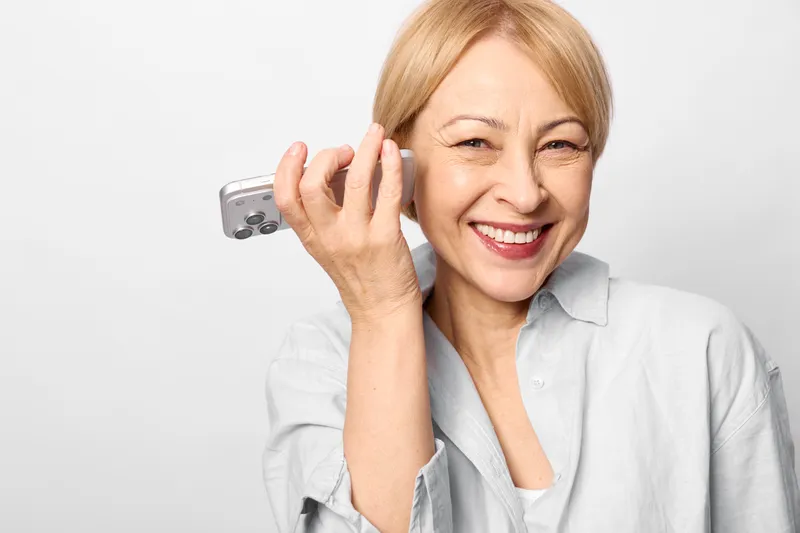 Smiling woman portrait indoors, elderly age, holding smartphone to ear, making a call, warm friendly expression, light gray shirt, clean white background, close up shot