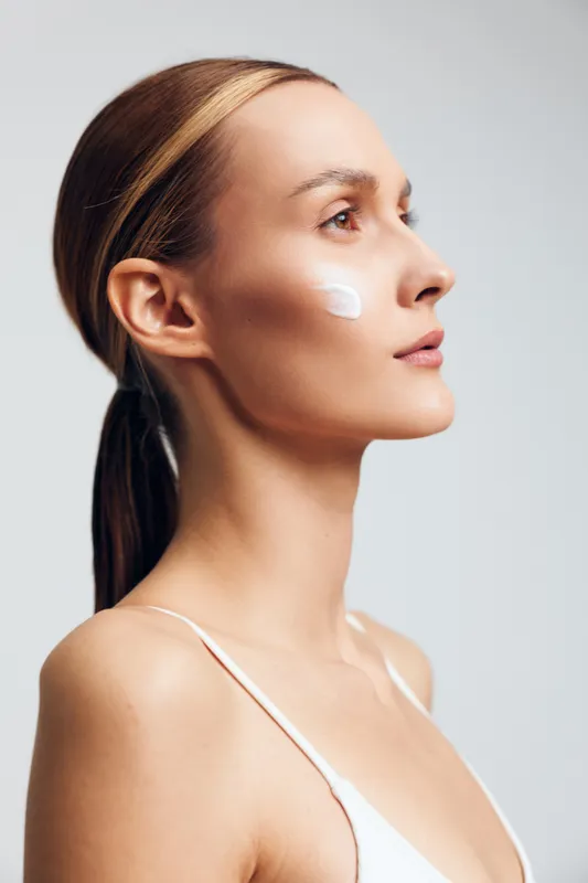 Smiling woman with light makeup applying cream on her cheek. Close up on a neutral background. Concept of skincare, self care, and wellness. Soft lighting creates a calm, fresh mood.