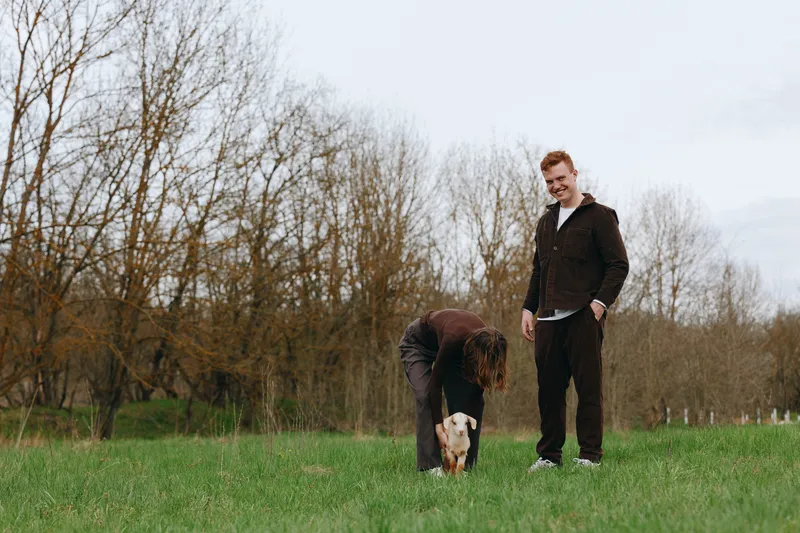 A man stands in a park while a friend crouches to interact with a small dog on the grass, creating a relaxed outdoor lifestyle moment under soft daylight and trees.