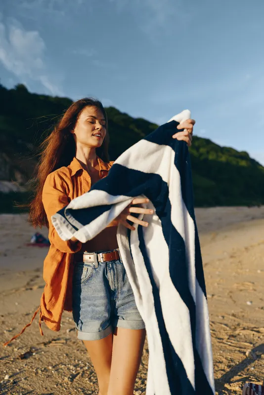Woman on beach holds striped blanket outdoors in summer sunlight, wearing casual denim shorts and jacket, scenic mountains and clouds behind, relaxed travel vacation mood, warm day at shoreline