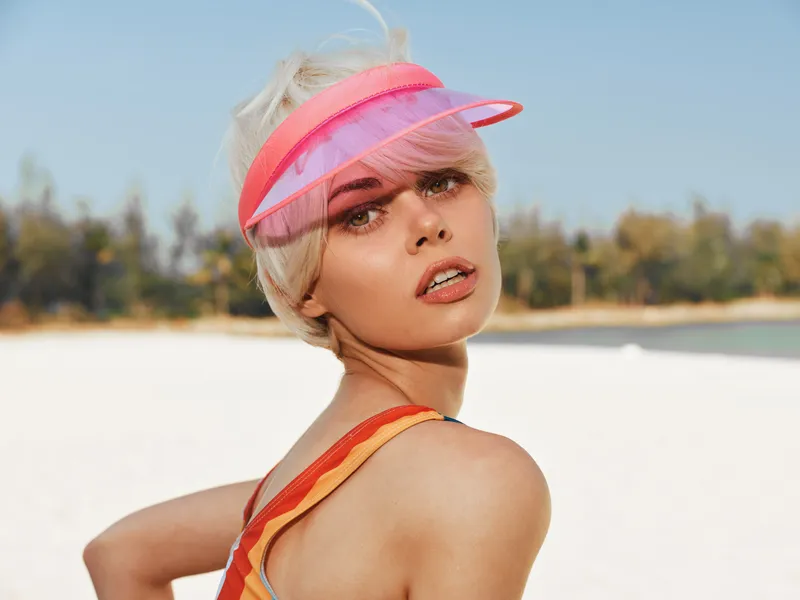 Portrait of a stylish woman on a sunny beach wearing a pink visor, orange strap swimsuit, and windswept hair, capturing bold summer charm and relaxed confidence.