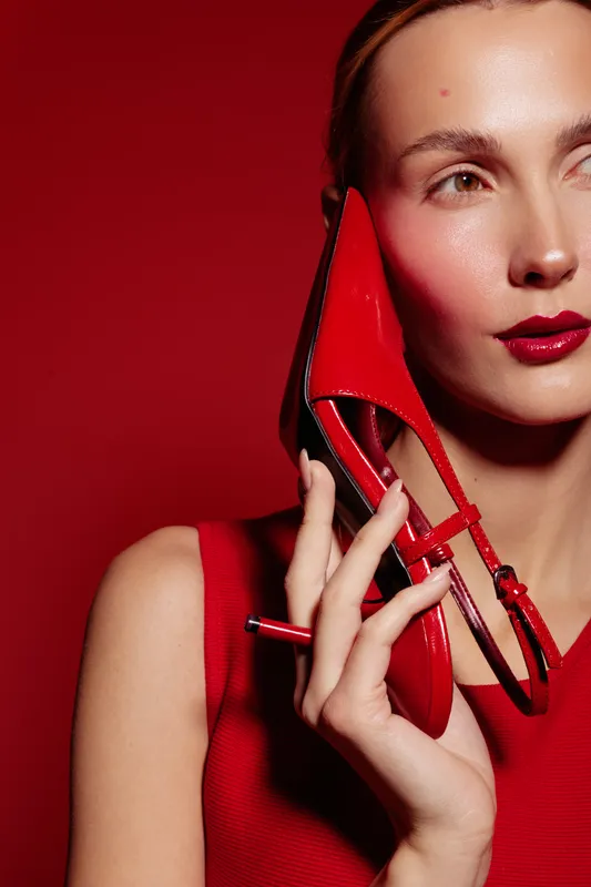 Close-up of a woman in red dress holding a red high heel shoe near her face on red background. Concept of fashion, style, elegance, and beauty with vibrant color contrast.