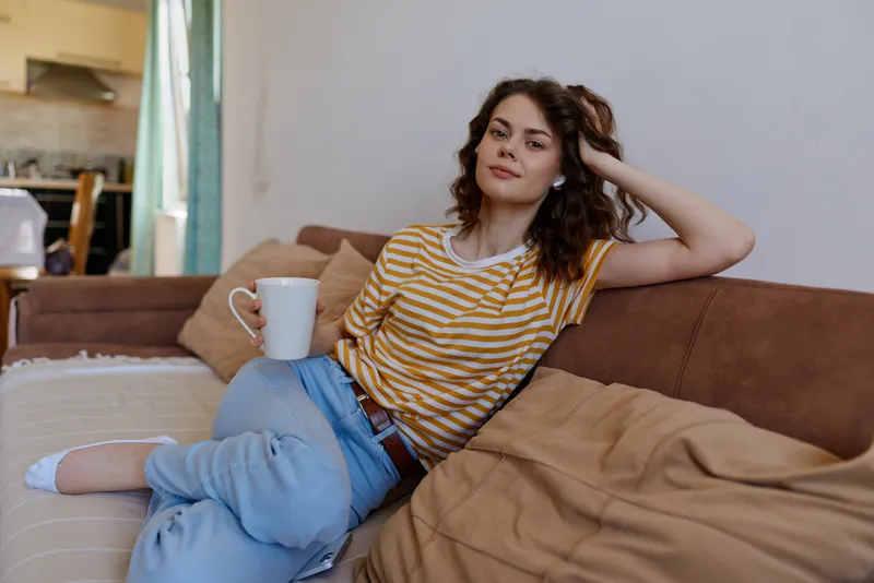 Casual woman at home, seated on a sofa with a mug, relaxed pose, striped tee and jeans, warm natural light, cozy interior, everyday comfort, lifestyle moment in a bright room with soft textiles and