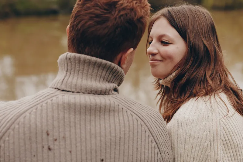 Smiling woman with brown hair in a knit sweater beside a calm river, sharing a quiet outdoor moment. Lifestyle mood focused on connection, nature, and comfort under soft natural light.