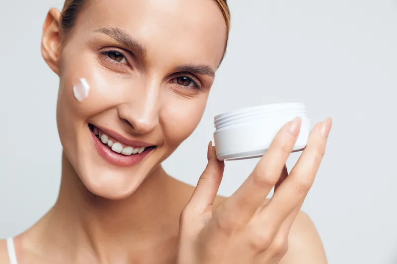 A smiling young woman with clear skin holds a white jar of moisturizer near her face, applying cream to her cheek. The image represents skincare, beauty, and self-care routines.