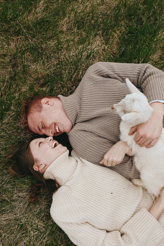 Joyful couple lying on grass in knit sweaters, embracing a white cat. Close, intimate moment highlighting affection, comfort, and relaxed lifestyle outdoors with soft natural light.