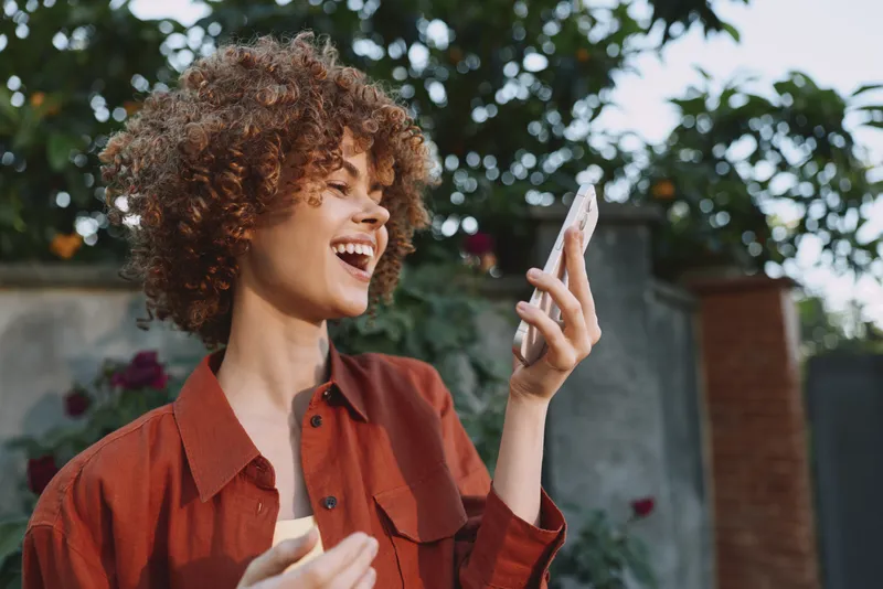 Joyful woman outdoors, smiling at her phone, casual red shirt, sunny day, natural light, relaxed pose, vibrant greenery backdrop