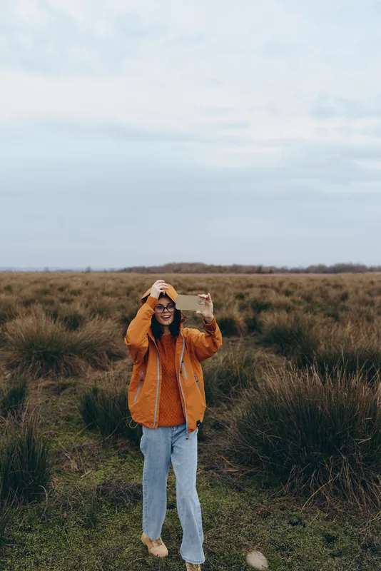 Woman lifestyle in an outdoor grassy field under cloudy sky, wearing orange jacket, beanie and sunglasses, holding a printed photo close to her face, calm relaxed mood.
