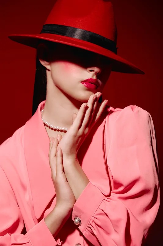 Woman wears a red hat, pink blazer, and pearl necklace against a red background combining fashion, beauty, and elegant creative style.