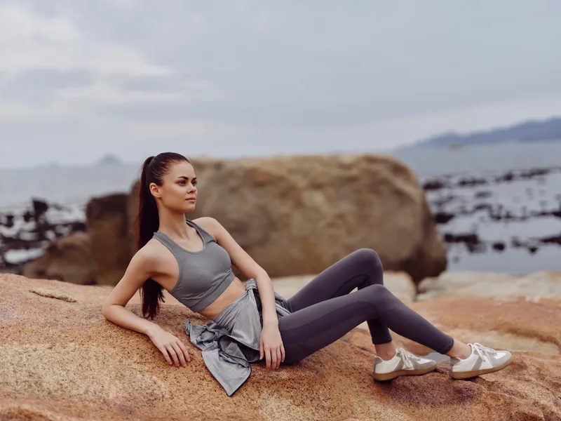 Young athletic woman in grey sportswear sitting on a rock by the sea, showcasing confidence and relaxation in a calm outdoor setting