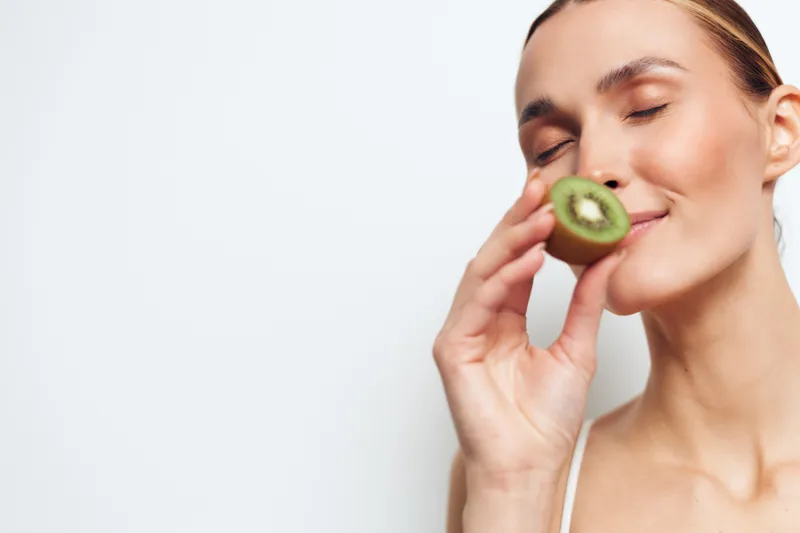 Young woman holding half of fresh kiwi fruit near face, eyes closed, enjoying aroma, natural skincare concept, soft lighting, minimalistic white background.