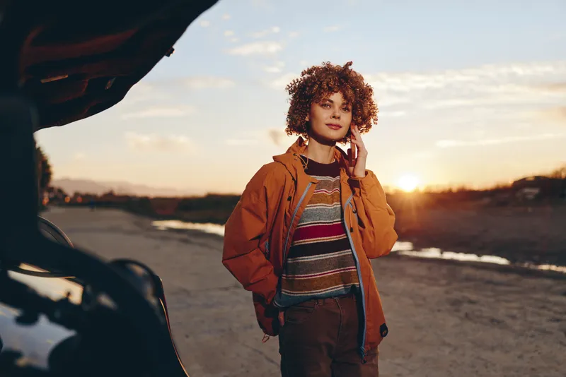 Curly haired woman lifestyle outdoors standing by open car door at sunset, warm golden light, relaxed smile, casual jacket and jeans, travel mood, natural landscape and soft breeze