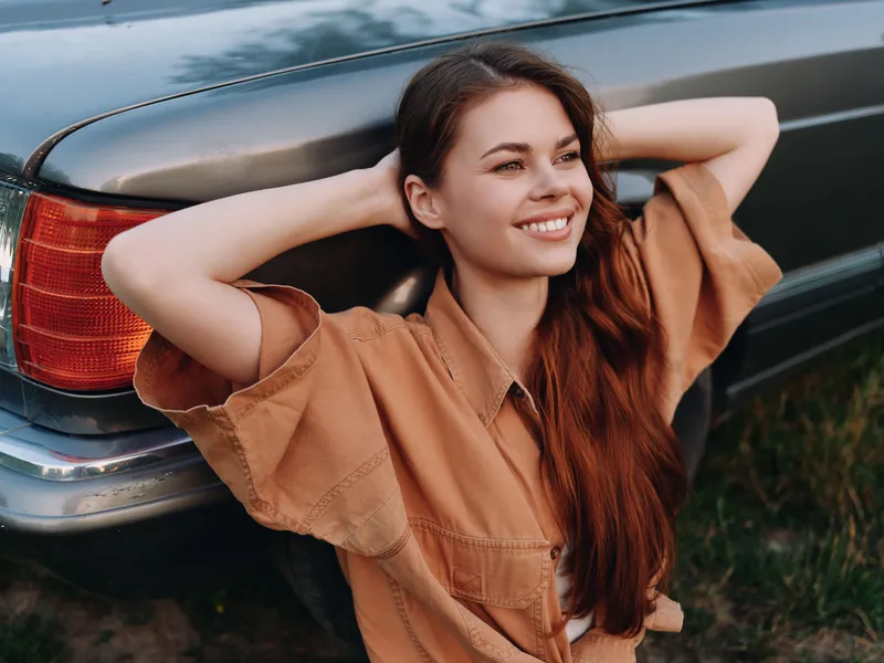 Young woman with long hair smiling outdoors, leaning against a vintage car, wearing a casual tan outfit, emanating a carefree and joyful vibe in natural light.