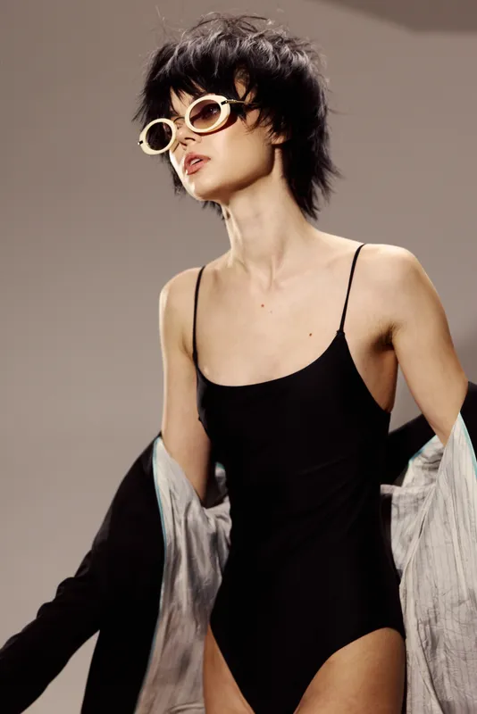 Stylish woman in black bodysuit wearing sunglasses with a trendy short haircut and minimalistic background.