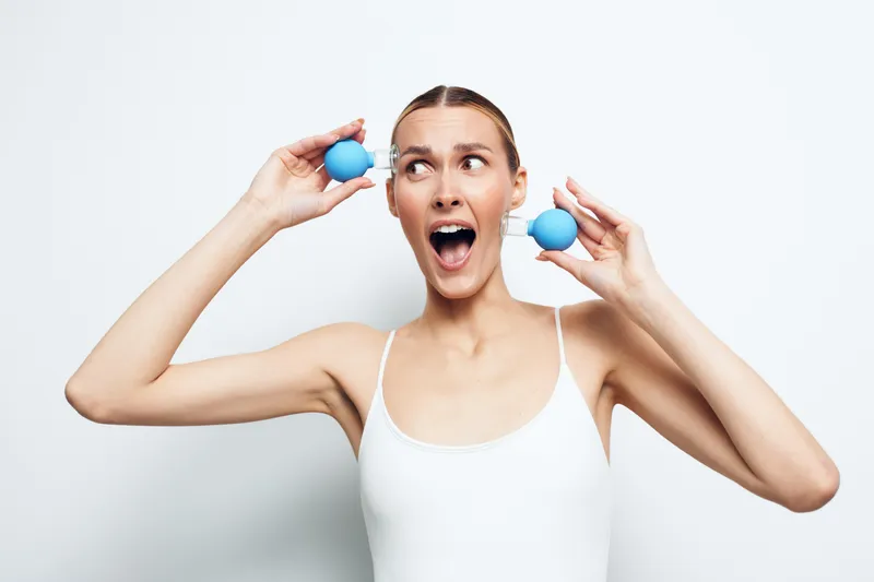 Young woman in white tank top holding two blue stress balls near her head with surprised expression on white background, concept of relaxation and mental health.
