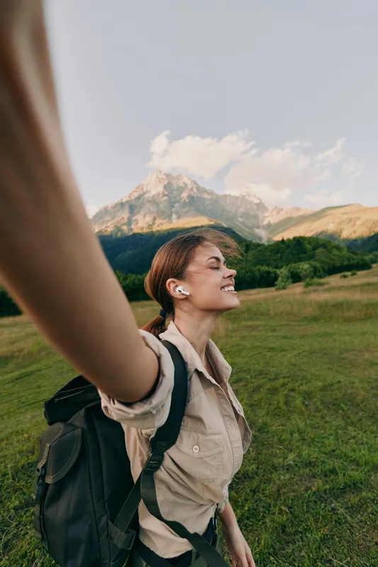 A smiling woman in a beige shirt with a backpack stands in a sunny meadow, arms raised. Alpine mountains rise behind, conveying freedom, adventure, and outdoor joy.