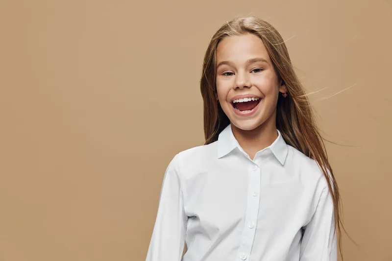 Girl with long hair and white shirt smiling happily on beige background. Portrait of cheerful child with open mouth and bright expression for childhood joy and positivity.
