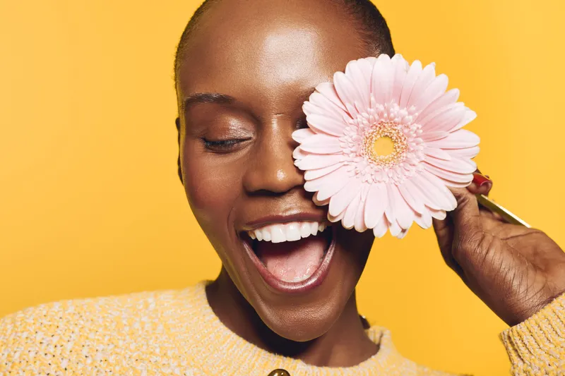 Happy African woman with dark skin laughs joyfully holding pink flower near eye. Yellow background and casual sweater create bright, cheerful mood. Concept for beauty, nature, and happiness.