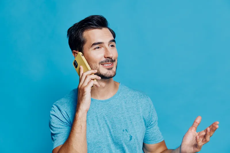 Smiling man wearing casual blue t-shirt talking on smartphone against solid bright blue background, presenting joyful attitude and lifestyle concept.