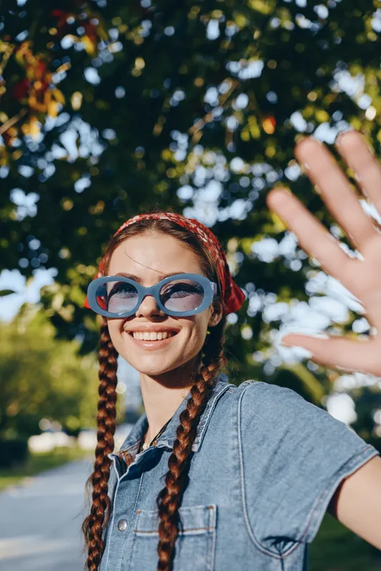 smiling woman with braids wearing blue sunglasses and denim shirt outdoors on a sunny day with greenery background