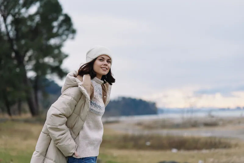 Winter outdoor portrait of a woman in a puffy coat and beanie by a lake, smiling as wind blows through the landscape, calm water and distant trees create a peaceful seasonal mood
