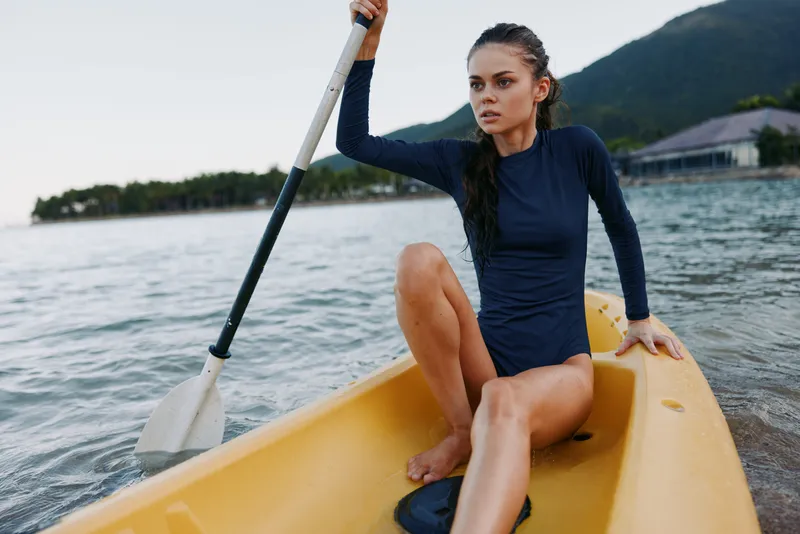 Athletic woman paddling a yellow kayak in calm water, wearing a navy long-sleeve swimsuit, wet hair, focused expression, sunlight on the lake, scenic mountain backdrop.