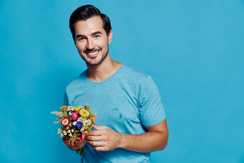 Smiling man holding a colorful bouquet of flowers dressed in a light blue t-shirt standing against a bright blue background. Floral lifestyle concept.