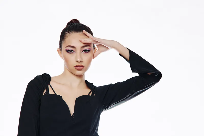 Fashion model with creative eye makeup poses with hand near forehead in a black modern outfit on white background. Stunning beauty concept with expressive vogue styling and attitude.
